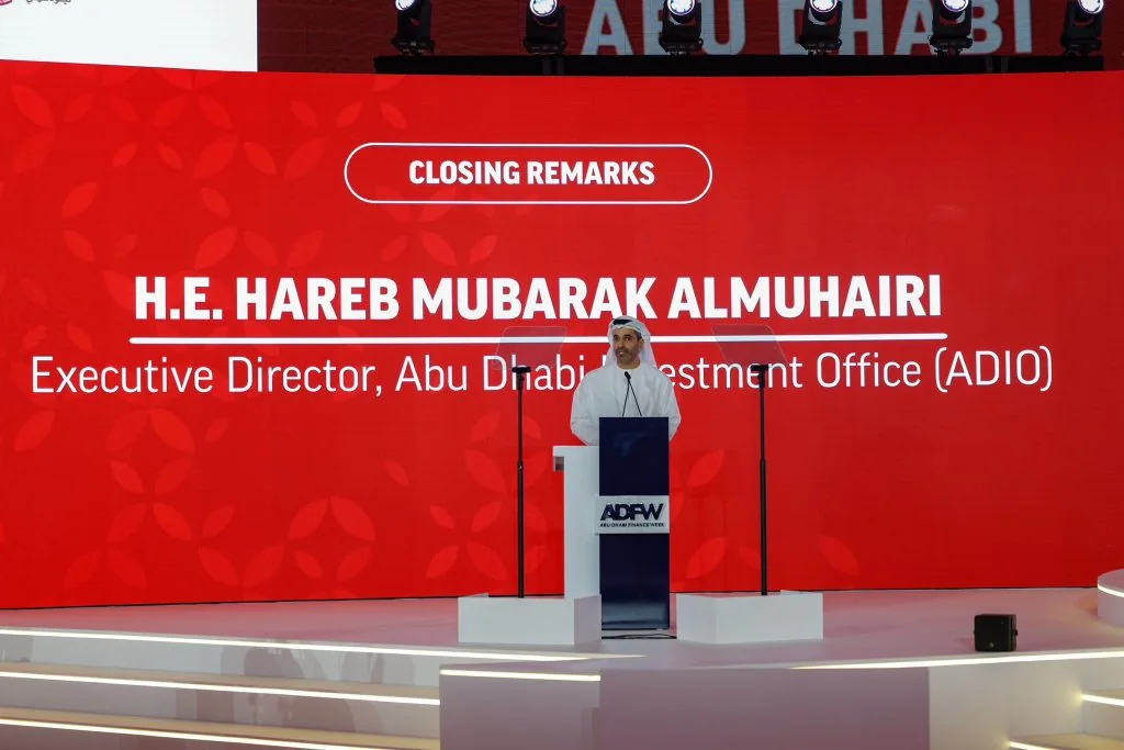 A speaker at a lectern in front of a red LED name card: a rounded “Closing Remarks” label above a name and title in white capitals.