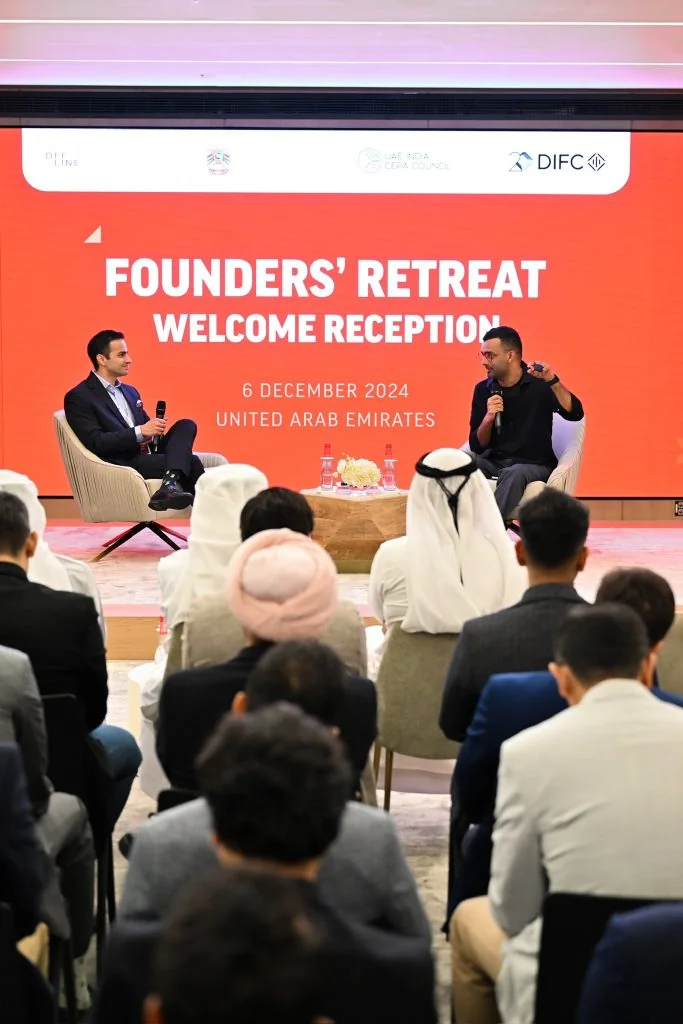 Two people in armchairs on a low stage, in front of a red screen reading “Founders’ Retreat Welcome Reception, 6 December 2024, United Arab Emirates”, with an audience in the foreground.
